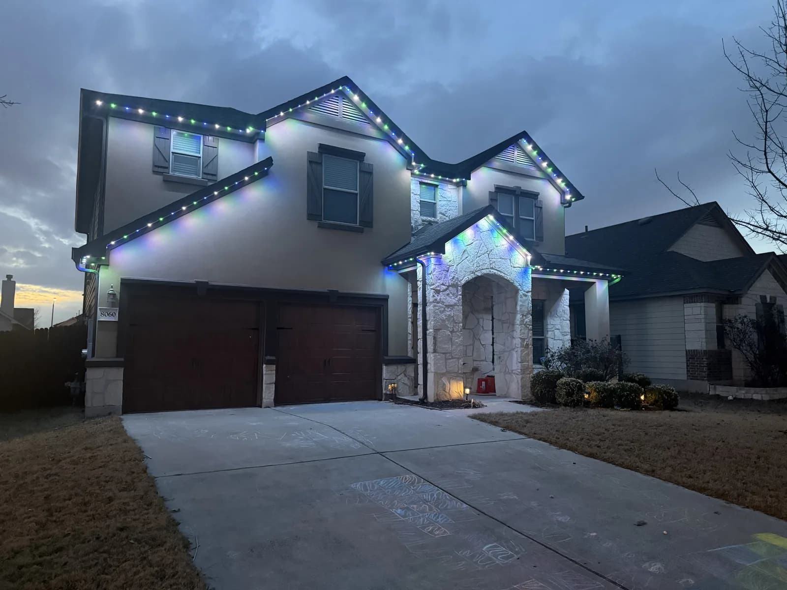 Blue, green, and white lights follow the upper roof and stone entry of a Round Rock home at dusk.